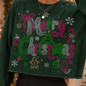 May include: A dark green long-sleeve cropped sweatshirt featuring the text Merry Christmas in a retro, multi-colored line art font. The design includes graphic illustrations of a Christmas tree, snowflakes, a candy cane, a gingerbread man, and ornaments.