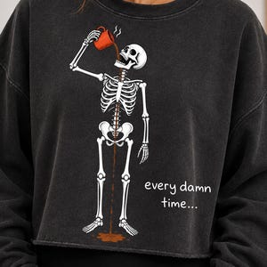 Charcoal gray folded t-shirt with a white skeleton pouring coffee from a red mug into its open mouth, coffee running down the spine, and the handwritten words every damn time, flat lay on a cream blanket with a straw hat. Funny skeleton coffee PNG shirt design.