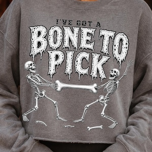 May include: A heather gray long-sleeve sweatshirt featuring a graphic of two skeletons pulling on a large bone. The text "I'VE GOT A BONE TO PICK" is printed in a distressed, white, dripping font above the skeletons. The garment has a crew neckline.