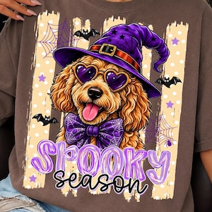 Puede incluir: Una camiseta marrón con el gráfico de un perro de color dorado que lleva un sombrero de bruja morado, gafas de sol en forma de corazón y una pajarita morada. El diseño incluye murciélagos, telarañas y el texto Spooky season en letras moradas.