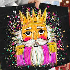 Puede incluir: Manta negra con una colorida ilustración de un cascanueces con corona dorada, bigote blanco y chaqueta rosa. El diseño incluye salpicaduras de pintura coloridas, creando una estética festiva y caprichosa.