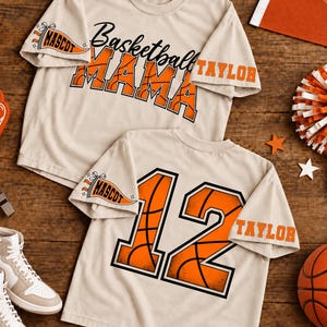 May include: Two beige t-shirts featuring orange basketball-themed graphics. One shirt displays the text Basketball MAMA with a pennant reading MASCOT and the name TAYLOR. The other shirt shows the number 12 with a pennant and the name TAYLOR.