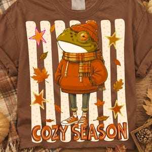 May include: A brown t-shirt featuring a graphic of a frog wearing an orange beanie, a plaid scarf, and an orange hoodie. The design includes vertical cream-colored stripes, scattered autumn leaves, stars, and the text COZY SEASON in orange.