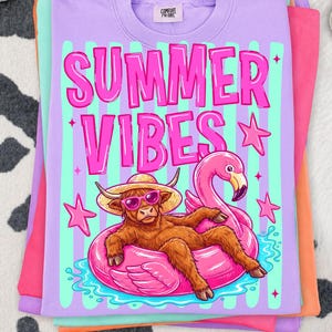 Op de afbeelding: Een stapel kleurrijke t-shirts. Het bovenste shirt is lavendelkleurig met de woorden "SUMMER VIBES" in roze, samen met een cartoon van een Schotse hooglander met een zonnebril en een flamingo-drijver. De shirts zijn gestapeld op een koeienprint achtergrond.