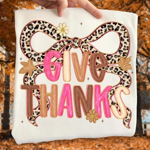 May include: Cream-colored t-shirt featuring the words "GIVE THANKS" in pink, brown, and tan letters. The text is surrounded by a leopard print bow and floral accents. The shirt is held up by a hand.