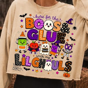 K&ouml;nnte beinhalten: Ein beiges, lang&auml;rmeliges, bauchfreies Sweatshirt mit einer bunten Halloween-Grafik und dem Text "here for the BOOS & GLUE with my LILGHOULS". Das Design zeigt Zeichentrickfiguren wie eine Klebeflasche, einen Apfel, ein Gespenst und eine Katze.