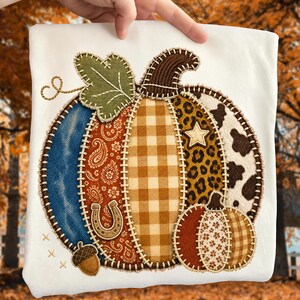 May include: A white t-shirt featuring a large pumpkin applique made of various fabric patches including blue denim, orange paisley, tan gingham, leopard print, and cow print. The design includes a green leaf, a horseshoe, and an acorn.