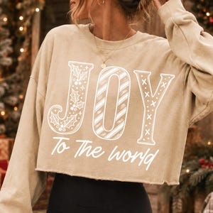 May include: A beige long-sleeve cropped sweatshirt featuring the white text JOY, where the J is decorated with holly leaves, the O has diagonal stripes, and the Y has an X pattern. Below, the white text reads To The world in script.