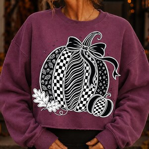 K&ouml;nnte beinhalten: Ein dunkelweinrotes Langarm-Sweatshirt mit einer gro&szlig;en Grafik von zwei K&uuml;rbissen. Der Hauptk&uuml;rbis hat Abschnitte mit Karomustern, Wellenlinien und Bl&auml;ttern, gekr&ouml;nt von einer schwarzen Schleife. Ein kleinerer karierter K&uuml;rbis steht daneben.