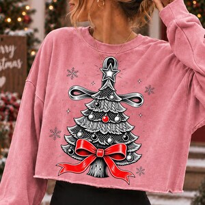 K&ouml;nnte beinhalten: Ein altrosa lang&auml;rmeliges bauchfreies Sweatshirt mit einem Grafikdruck eines stilisierten Weihnachtsbaums. Der Baum ist in Schwarz-Wei&szlig; gehalten, mit einer leuchtend roten Schleife an der Basis, einer roten Kugel und einem wei&szlig;en Stern an der Spitze, umgeben von Schneeflocken.
