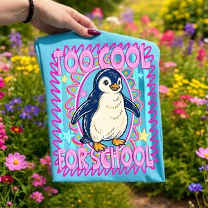 K&ouml;nnte beinhalten: Hellblaues T-Shirt mit einem Cartoon-Pinguin-Design. Der Pinguin hat einen wei&szlig;en Bauch und blaue Fl&uuml;gel. Der Text "TOO COOL FOR SCHOOL" ist rosa mit wei&szlig;er Umrandung, und das Design enth&auml;lt Sterne und einen wellenf&ouml;rmigen Rand.
