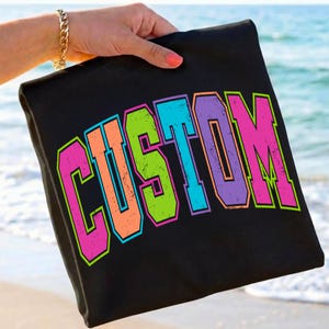 Pu&ograve; includere: Borsa tote nera con la scritta "CUSTOM" in un carattere colorato e invecchiato in stile collegiale. Il testo presenta colori rosa neon, arancione, verde, blu e viola con un contorno verde neon. La borsa &egrave; tenuta su uno sfondo di spiaggia.