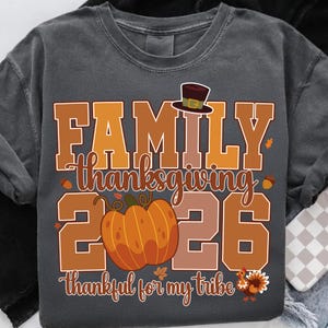 Pode incluir: Uma t-shirt cinzento-carv&atilde;o com um gr&aacute;fico que inclui o texto FAMILY THANKSGIVING 2026 e thankful for my tribe. O design apresenta um chap&eacute;u de peregrino castanho, uma ab&oacute;bora laranja, bolotas, folhas de outono e uma ilustra&ccedil;&atilde;o de um peru.