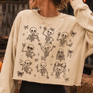 Op de afbeelding: Een beige cropped sweatshirt met lange mouwen en een zwarte grafische print van meerdere skeletten in verschillende houdingen, waaronder hartjeshanden, vredestekens en strikjes, afgewisseld met kleine ster- en striksymbolen.