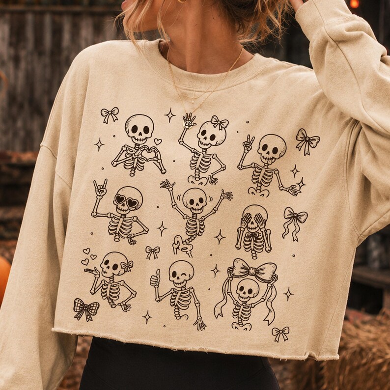 Op de afbeelding: Een beige cropped sweatshirt met lange mouwen en een zwarte grafische print van meerdere skeletten in verschillende houdingen, waaronder hartjeshanden, vredestekens en strikjes, afgewisseld met kleine ster- en striksymbolen.
