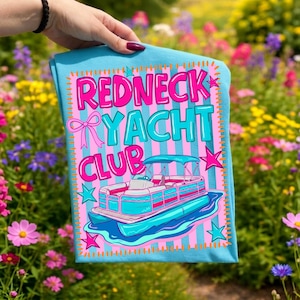 May include: A light blue t-shirt with the words "REDNECK YACHT CLUB" in pink and turquoise, featuring a cartoon pontoon boat. The design includes pink stars and a bow, set against a striped background.