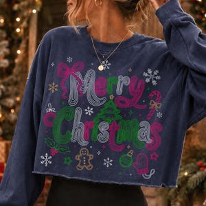 May include: A navy blue long-sleeve cropped sweatshirt featuring the text Merry Christmas in a wavy, multi-colored font. The design includes graphic illustrations of a Christmas tree, a gingerbread man, snowflakes, a candy cane, and a mitten.