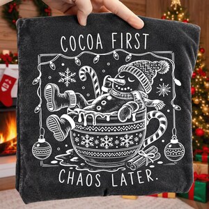 Op de afbeelding: Een antracietgrijs T-shirt met een witte afbeelding van een sneeuwpop die ontspant in een grote mok. De tekst luidt COCOA FIRST CHAOS LATER. Het ontwerp bevat een zuurstok, sneeuwvlokken, kerstverlichting en kerstballen in witte lijntekening.