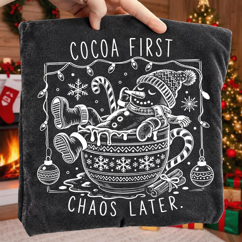 Op de afbeelding: Een antracietgrijs T-shirt met een witte afbeelding van een sneeuwpop die ontspant in een grote mok. De tekst luidt COCOA FIRST CHAOS LATER. Het ontwerp bevat een zuurstok, sneeuwvlokken, kerstverlichting en kerstballen in witte lijntekening.