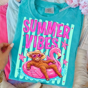 Może przedstawiać: Turkusowa koszulka Comfort Colors z grafiką z napisem "SUMMER VIBES". Grafika przedstawia brązową krowę Highland w okularach przeciwsłonecznych i słomkowym kapeluszu, relaksującą się na r&oacute;żowym pływaku z flamingiem. R&oacute;żowe gwiazdki i paski dopełniają projekt.