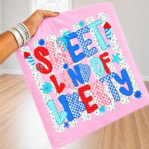 May include: A pink fabric square featuring the text SWEET LAND OF LIBERTY in a mix of red, blue, and light blue checkered and polka dot patterns. The design includes small firework illustrations and blue flowers on a white background with black dots.