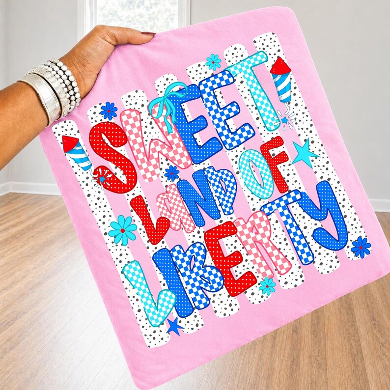 May include: A pink fabric square featuring the text SWEET LAND OF LIBERTY in a mix of red, blue, and light blue checkered and polka dot patterns. The design includes small firework illustrations and blue flowers on a white background with black dots.