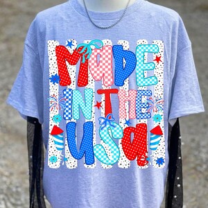 Pode incluir: Uma t-shirt cinza-claro com uma estampa gr&aacute;fica com o texto MADE IN THE USA. O design inclui padr&otilde;es em vermelho, branco e azul com bolinhas, xadrez e estrelas, acentuados por ilustra&ccedil;&otilde;es de fogos de artif&iacute;cio e pequenos la&ccedil;os azuis.