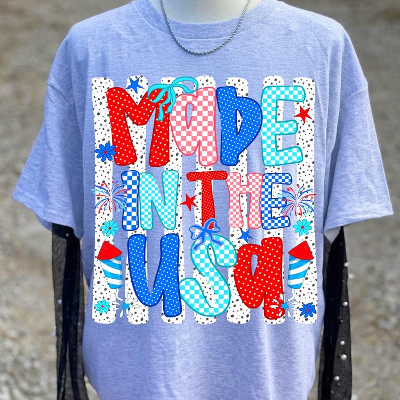 Pode incluir: Uma t-shirt cinza-claro com uma estampa gr&aacute;fica com o texto MADE IN THE USA. O design inclui padr&otilde;es em vermelho, branco e azul com bolinhas, xadrez e estrelas, acentuados por ilustra&ccedil;&otilde;es de fogos de artif&iacute;cio e pequenos la&ccedil;os azuis.