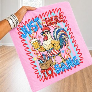 May include: Pink blanket with a cartoon rooster wearing sunglasses and patriotic shorts, holding a beer mug. The blanket has the text "JUST HERE TO BANG" in a wavy design. The rooster is standing next to a wooden crate of fireworks.