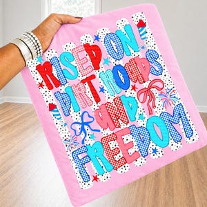 Op de afbeelding: Een roze vierkant stoffen item met de tekst "RISED ON DIRT ROADS AND FREEDOM" in rood en blauw geblokte en effen lettertypes. Het ontwerp bevat zwarte stippen, blauwe en rode vuurwerkillustraties en blauwe en roze lintstrikken.