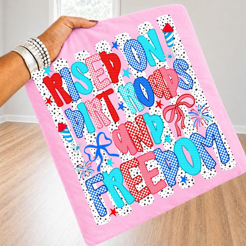 Op de afbeelding: Een roze vierkant stoffen item met de tekst "RISED ON DIRT ROADS AND FREEDOM" in rood en blauw geblokte en effen lettertypes. Het ontwerp bevat zwarte stippen, blauwe en rode vuurwerkillustraties en blauwe en roze lintstrikken.