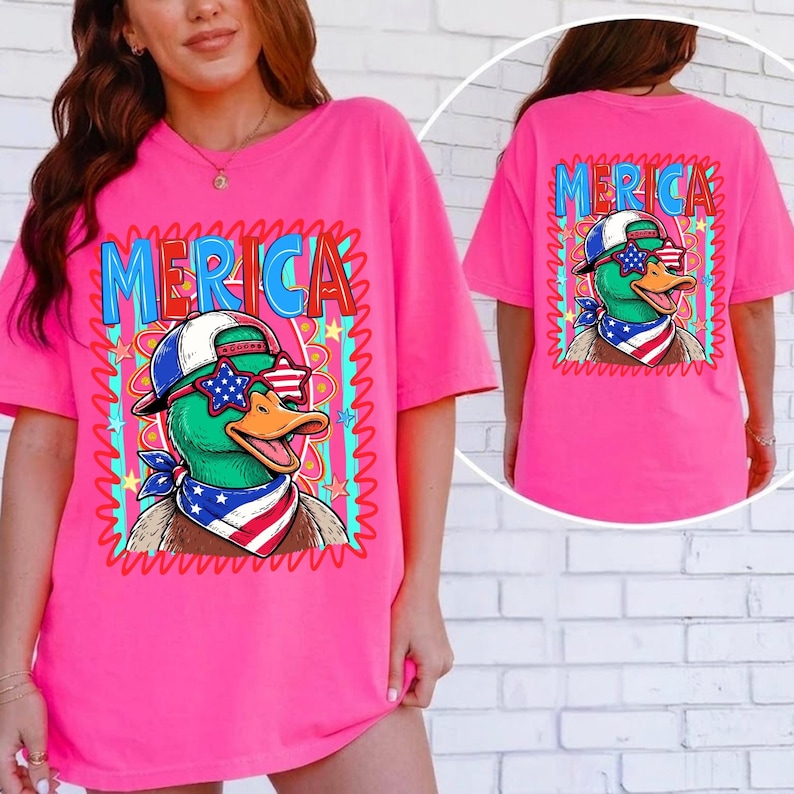 May include: Bright pink t-shirt featuring a cartoon duck wearing sunglasses, a bandana, and a baseball cap with the word "MERICA" in a colorful design. The same design is on the back of the shirt.