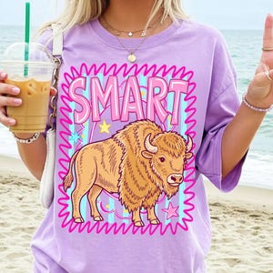 May include: A light purple t-shirt featuring a cartoon bison illustration with the word "SMART" in pink letters. The bison is tan with a fluffy mane and horns. The design includes stars and wavy lines.