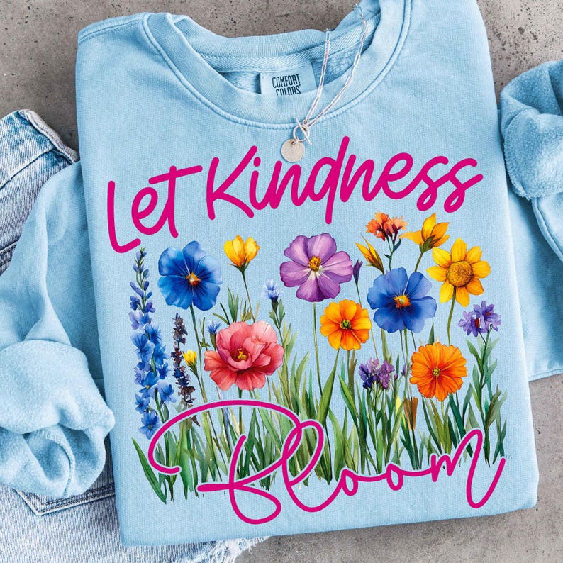 May include: A light blue sweatshirt with the words "Let Kindness Bloom" in pink script. The design features a colorful floral arrangement with blue, yellow, orange, and purple flowers. A silver necklace is visible.
