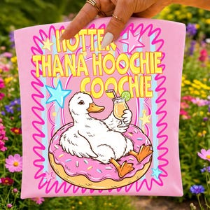 Może przedstawiać: R&oacute;żowa koszulka z ż&oacute;łtym napisem "Hotter Than a Hoochie Coochie". Koszulka przedstawia kresk&oacute;wkową kaczkę relaksującą się na pączku i trzymającą koktajl. Projekt zawiera gwiazdy i faliste linie.