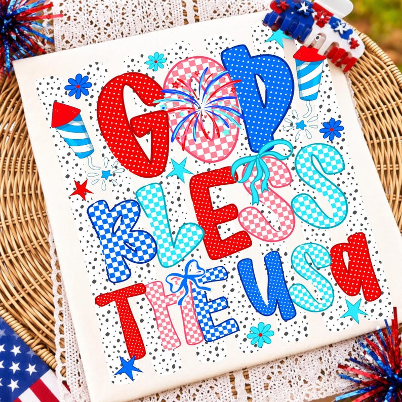 May include: A white rectangular sign featuring the text GOD BLESS THE USA in red, blue, and pink checkered and polka-dot patterns. The design includes illustrations of fireworks, stars, and flowers on a background of small black dots.