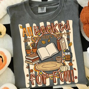 K&ouml;nnte beinhalten: Ein anthrazitfarbenes T-Shirt mit einem Grafikdruck und dem Text "All Booked Just one More chapter for Fall". Das Design zeigt ein offenes Buch, einen B&uuml;cherstapel, eine Kaffeetasse, eine French Press, Topfpflanzen und einen K&uuml;rbis mit Herbstbl&auml;ttern.