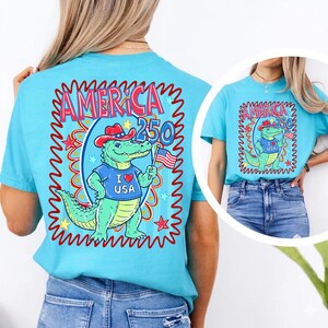 Puede incluir: Una camiseta azul claro con un dise&ntilde;o patri&oacute;tico. El gr&aacute;fico incluye el texto "AMERICA 250" y un cocodrilo de dibujos animados con un sombrero de vaquero y una bandera estadounidense. El cocodrilo lleva una camiseta azul que dice "I ❤️ USA".