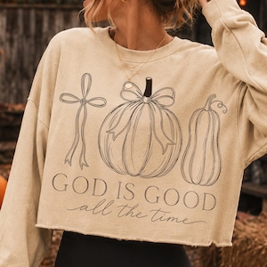 K&ouml;nnte beinhalten: Ein beiges, lang&auml;rmeliges, bauchfreies Sweatshirt mit einer schwarzen Strichgrafik, die ein Kreuz mit Schleife, einen K&uuml;rbis mit Schleife und einen Flaschenk&uuml;rbis zeigt. Der Text "GOD IS GOOD all the time" ist darunter in Schwarz aufgedruckt.