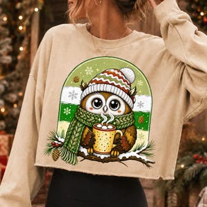 May include: A beige cropped sweatshirt featuring a graphic of an owl wearing a red, white, and green striped knit hat and a green scarf. The owl holds a yellow mug with white polka dots, sitting on a snowy branch with pine cones and snowflakes.