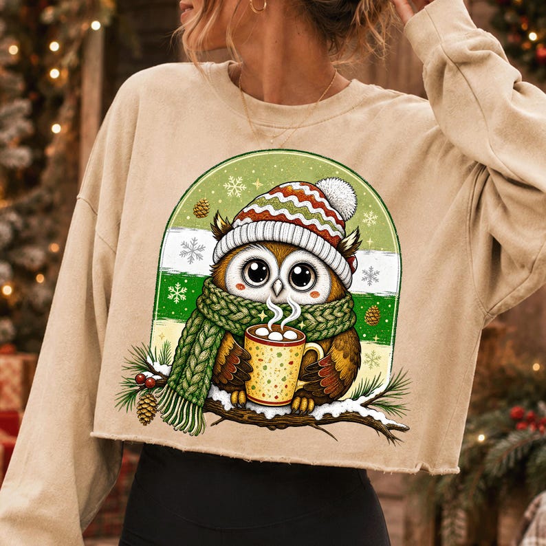 May include: A beige cropped sweatshirt featuring a graphic of an owl wearing a red, white, and green striped knit hat and a green scarf. The owl holds a yellow mug with white polka dots, sitting on a snowy branch with pine cones and snowflakes.
