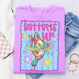 May include: A lilac sweatshirt featuring a colourful duck diving into water, with the words "BOTTOMS UP" in pink. The design includes stars and a striped background in blue and pink.