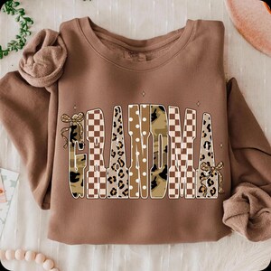 May include: A brown crewneck sweatshirt featuring the word GRANDMA printed in large letters. The letters have varied patterns including brown and white checkerboard, leopard print, camouflage, and white polka dots, with small bows on the G and A.
