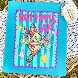 May include: A teal canvas tote bag with a vibrant illustration of a duck diving into water, surrounded by stars. The words "Bottoms Up" are written in pink above the duck. The bag has a pink scalloped border.