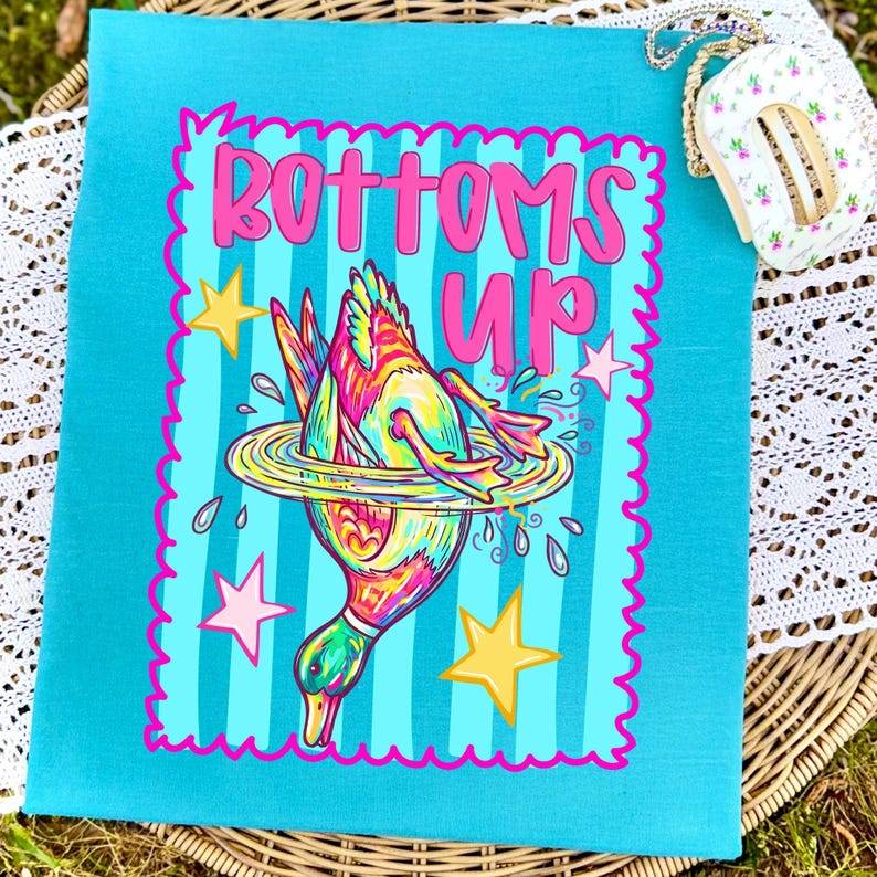 May include: A teal canvas tote bag with a vibrant illustration of a duck diving into water, surrounded by stars. The words "Bottoms Up" are written in pink above the duck. The bag has a pink scalloped border.