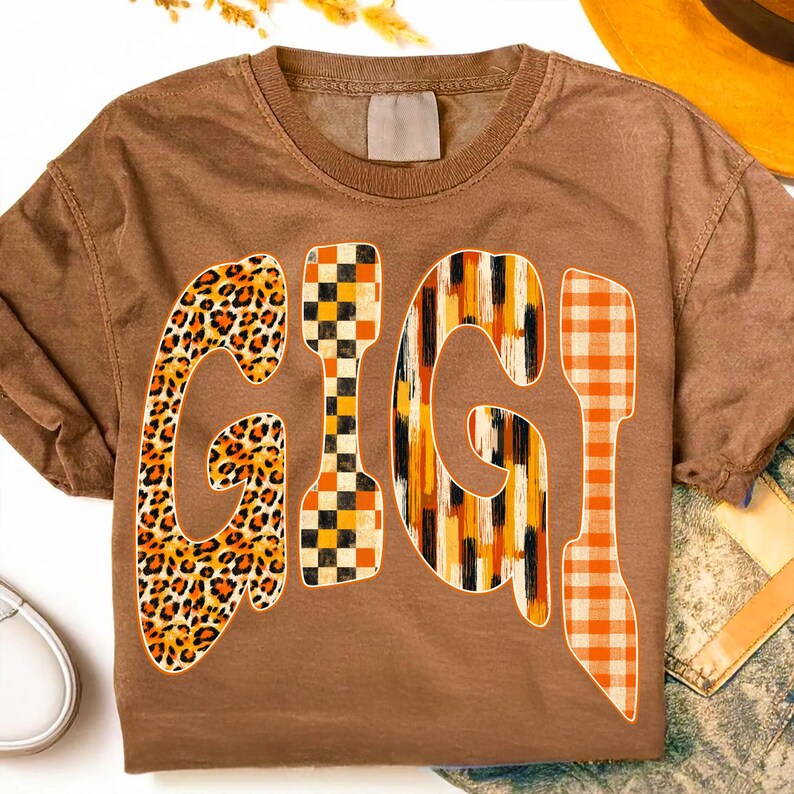 May include: A brown short-sleeve t-shirt featuring the word GIGI printed in large letters across the chest. Each letter has a distinct pattern: the first G is leopard print, the I is a black and orange checkerboard, the second G is vertical stripes, and the final I is orange plaid.