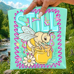 Op de afbeelding: Een lichtblauwe draagtas met een cartoonbij, een honingpot en het woord "STILL" in een turquoise bubbellettertype. Het ontwerp bevat roze sterren, bloemen en een golvende rand. De tas wordt door een hand omhoog gehouden.