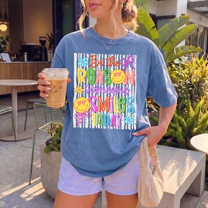 May include: A blue t-shirt with the text "Be the reason someone smiles today" in colorful letters. The shirt has a relaxed fit and is paired with striped shorts. A clear plastic cup with a brown beverage is held in the left hand.