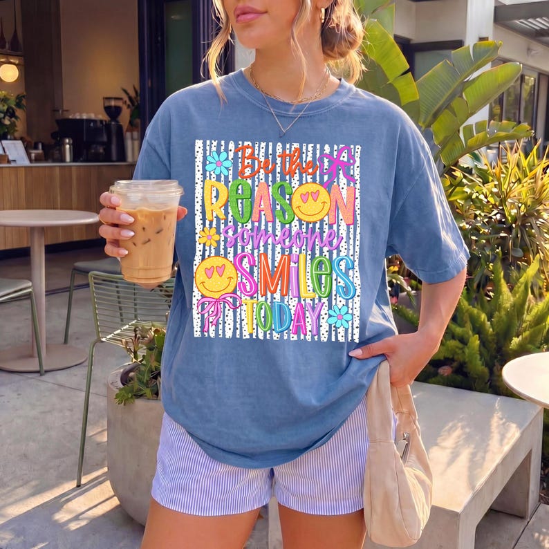 May include: A blue t-shirt with the text "Be the reason someone smiles today" in colorful letters. The shirt has a relaxed fit and is paired with striped shorts. A clear plastic cup with a brown beverage is held in the left hand.