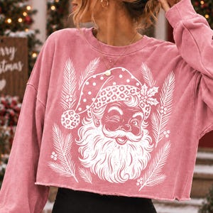Puede incluir: Una sudadera corta de manga larga en color rosa empolvado con un estampado gr&aacute;fico blanco de un Santa Claus gui&ntilde;ando un ojo, con un gorro de lunares y estampado de leopardo, rodeado de ramas de pino y bayas de acebo estilizadas.